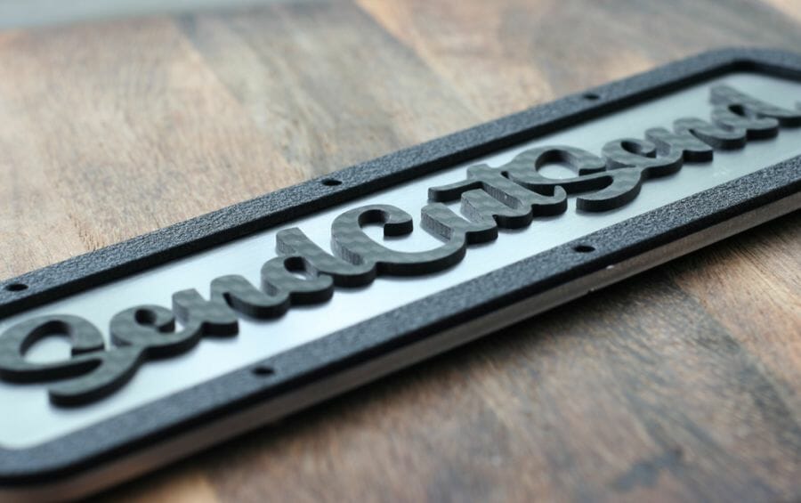 Image of a laser cut sheet metal part with the SendCutSend logo fixtured to it and cut out of carbon fiber